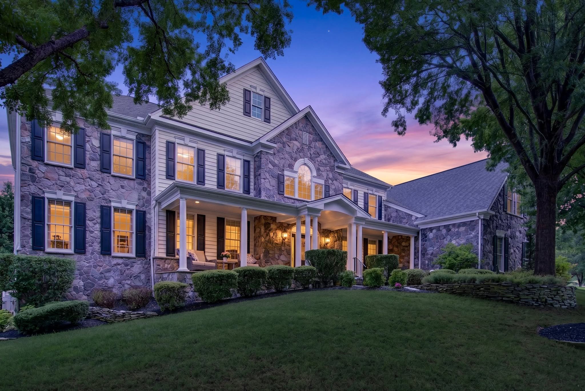 Stone colonial home with a lit front porch and warm window light photographed at twilight